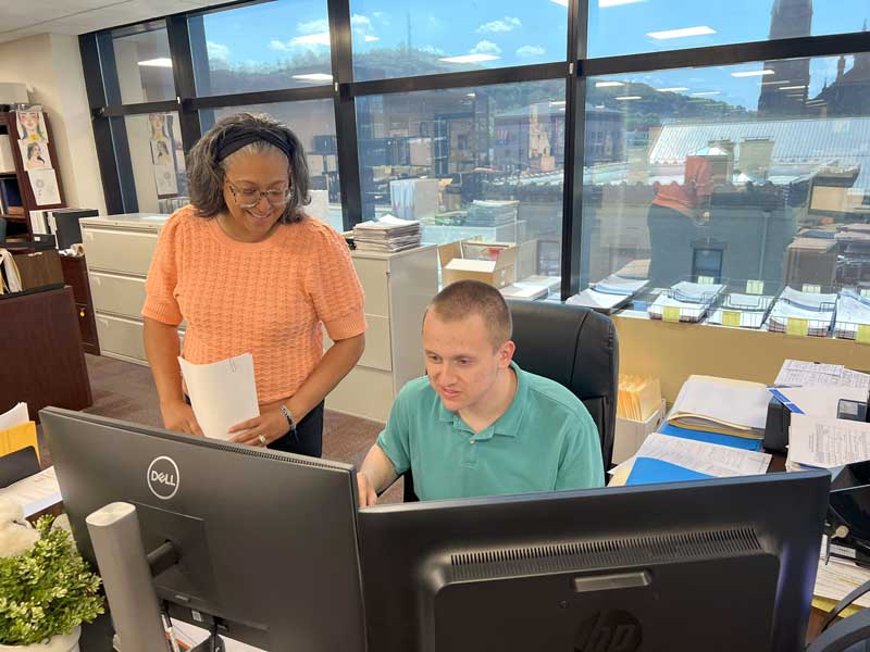 Peter HR Clerk - Passaic County Human Resources Department. Peter is seen working at a desk with another person standing to his right. Both are looking towards the dual monitors.