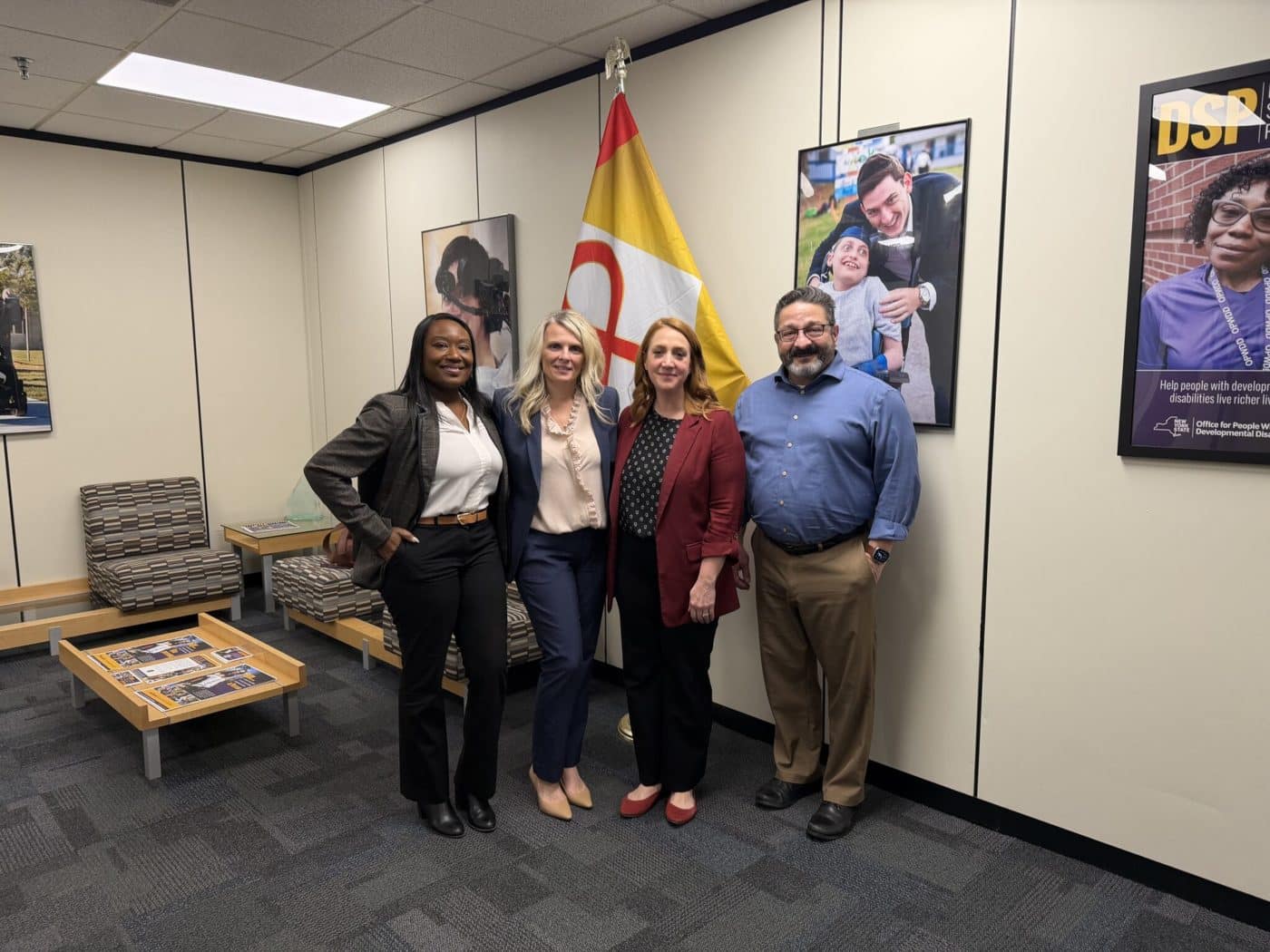 Fatimat Adekoya, Svetlana Repic-Qira, Willow Baer, and John Barbuto stand together in an office with New York’s red and gold neurodiversity flag displayed behind them.
