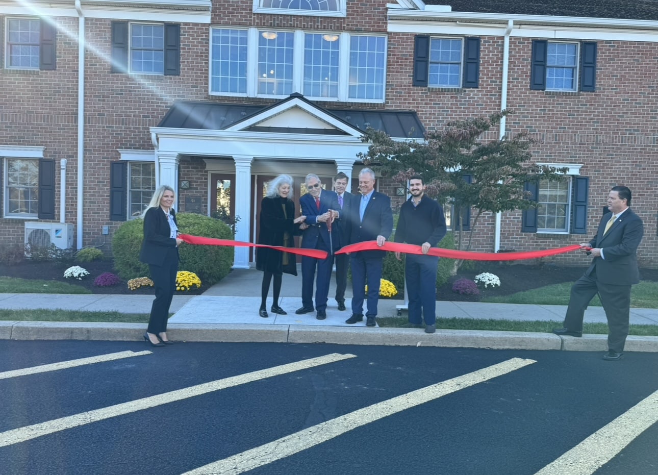 A group of officials and donors stands together during a ribbon-cutting ceremony as Philip E. Lian cuts the ribbon with oversized ceremonial scissors outside the Daily Plan It in Newtown, Pennsylvania.