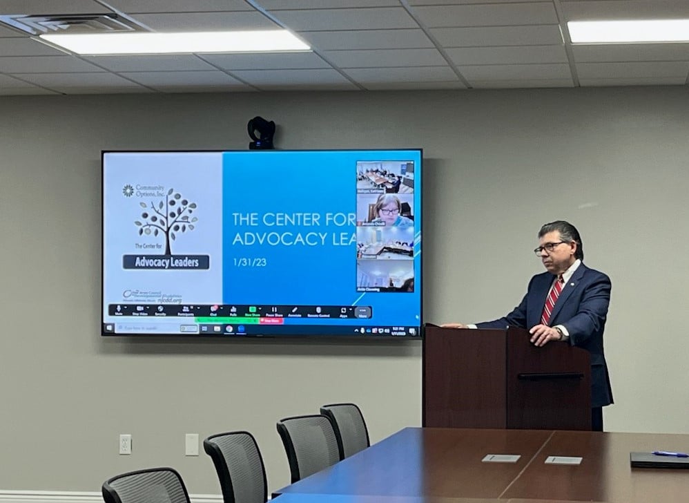 New Jersey State Senator Anthony Bucco standing in front of a projector screen displaying remote participants during a hybrid meeting.