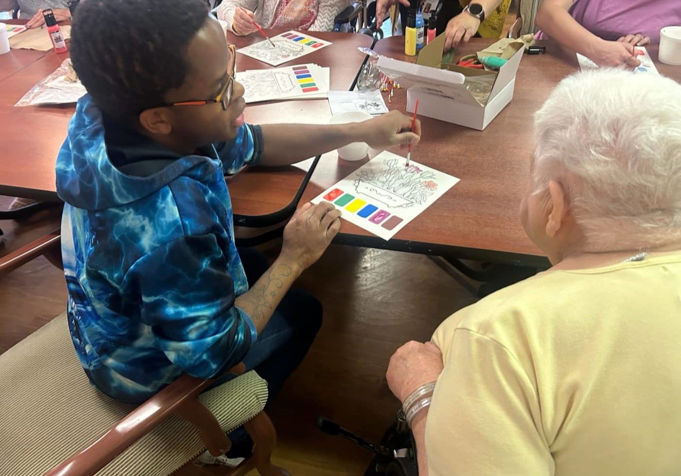 Ricky seated at a large table during a watercolor activity while volunteering at a nursing center, with other participants working on paintings nearby.